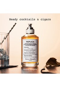 Flacon de parfum Maison Margiela Replica étiqueté Heady cocktails and cigars, verre avec agitateur et feuilles de cigare séchées sur la table.
