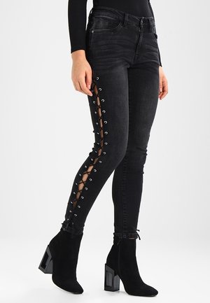 Femme portant un jean skinny noir avec des détails lacés sur les côtés extérieurs et des bottines noires à talons hauts avec talons géométriques.