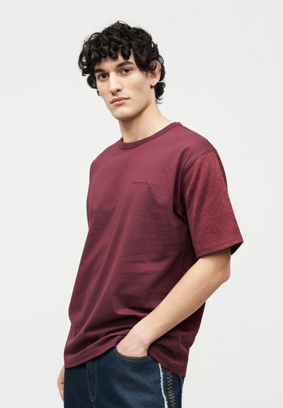 Νεαρός άνδρας με σγουρά σκούρα μαλλιά φοράει ένα μπορντό κοντομάνικο μπλουζάκι και σκούρο τζιν, στέκεται με το ένα χέρι στην τσέπη μπροστά σε ένα απλό φόντο.