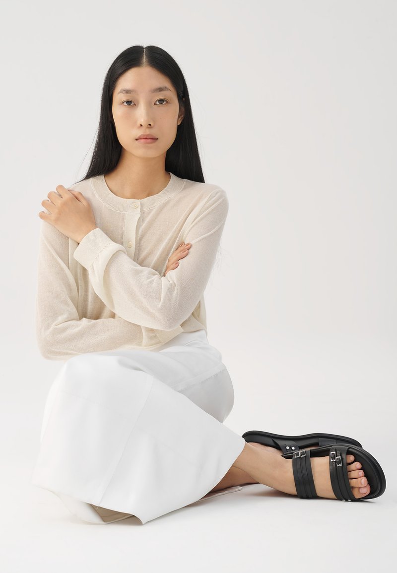 Hellbeige Bluse mit langen Ärmeln und Knöpfen, kombiniert mit einem weißen Rock; das Model trägt schwarze Riemchensandaletten und sitzt mit verschränkten Armen.