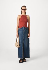 Haut en tricot rouge à bretelles, jupe maxi en denim bleu taille haute avec fente avant, tongs noires et sac à main beige ; lunettes de soleil rectangulaires.