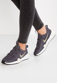 Mörklila sportskor med meshovandel, snörning och en gyllen färgad swoosh-logotyp; vit mellansula och svart yttersula; texturerad yta.