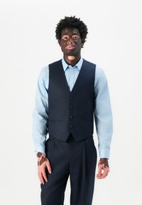 Mann mit Vitiligo, der ein hellblaues Hemd sowie eine dunkelblaue Weste und Hose trägt und vor einem weißen Hintergrund steht.