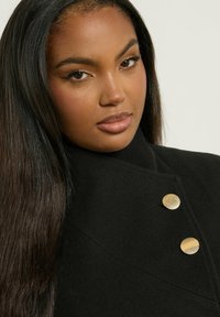 Abrigo de lana negro con un cuello alto, que presenta dos botones de oro texturizados y un diseño elegante y entallado. Textura suave, costuras mínimas.