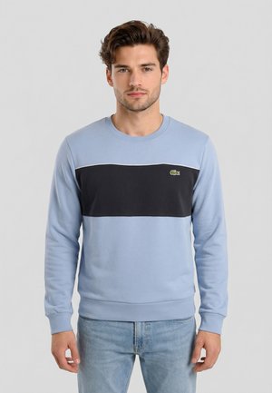 Uomo con capelli corti e mossi che indossa una felpa azzurro chiaro con una fascia orizzontale nera sul petto e jeans azzurri, rivolto in avanti.