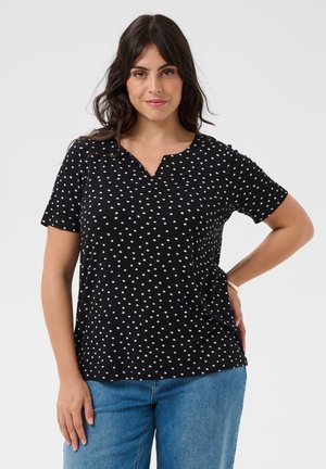 Vrouw met lang donker haar, gekleed in een zwart polkadot shirt met korte mouwen en blauwe spijkerbroek, staand met één hand op de heup tegen een witte achtergrond.