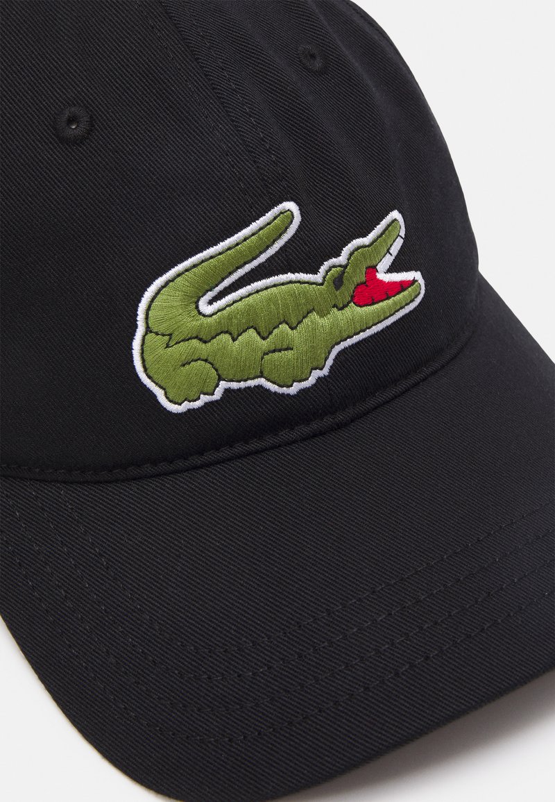 Gorra negra con visera curvada, que presenta un logotipo de cocodrilo verde bordado con un detalle rojo. Hecha de una tela texturizada con costuras cosidas.