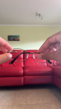 Metal eyeglasses with a thin silver frame, black plastic temples, and clear lenses, held in hand against a red sofa backdrop.