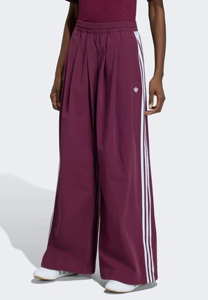 Personne portant un pantalon large bordeaux avec des rayures latérales blanches et des baskets blanches, debout devant un fond clair.