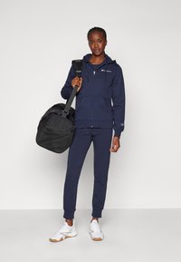 Marineblå hettegensere med glidelås og snorer, matchende joggebukser og hvite joggesko. Holder en svart duffelbag med strukturert bunn.