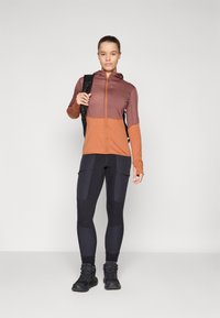 Hooded Sporttop in braun und orange mit Farbblockierung, langen Ärmeln, kombiniert mit schwarzen Leggings mit aufgesetzten Taschen und Wanderstiefeln.