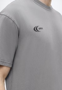 Ανοιχτό γκρι κοντομάνικη μπλούζα με μαύρο λογότυπο ARKK στο αριστερό στήθος, που φοριέται από άτομο που εμφανίζεται από τον λαιμό μέχρι τη μέση.