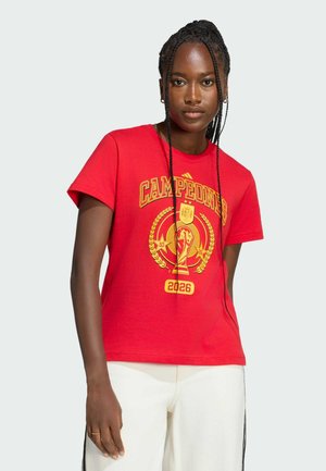 Femme aux cheveux tressés portant un t-shirt rouge "Campeones 2026" et un pantalon blanc, debout devant un fond clair uni.