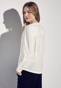 Vit långärmad blus med volangkrage, texturerade manschetter och sammannehmen detalj bak. Mjuk tyg med en slät yta.