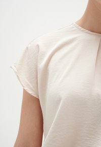 Lys beige top med korte, oppbrettede ærmer. Blødt, let tekstureret stof og rund halsudskæring. Minimalistisk design uden udsmykninger.