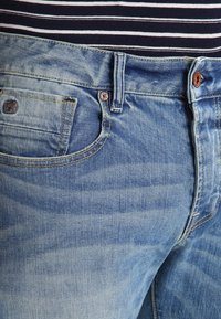 Ljusblå denimjeans med en distressed yta, femfickorsdesign och kontrasterande sömmar. Har en märkesknapp och nitar som dekoration.