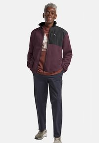 Veste en polaire bordeaux avec un panneau d'épaule noir, une fermeture éclair et un col montant ; associée à un sweat-shirt graphique marron et un pantalon bleu marine.