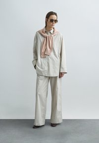 Chemise oversize beige à rayures, pantalon large assorti, pull rose drapé autour du cou, lunettes de soleil foncées, chaussures marron, fond minimaliste.