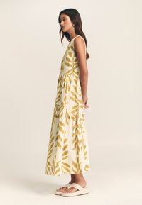 Dolga obleka v kremni barvi z rumenim vzorcem listov, brez rokavov, večplastnim zgornjim delom in sproščenim krojem. Nosijo jo s kremnimi platformnimi sandali.