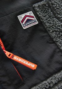 Tessuto esterno nero con pannello in pile grigio testurizzato. Presenta una toppa con il logo "Superdry Mountain Sport" blu, rosso e bianco e un cursore della cerniera arancione.