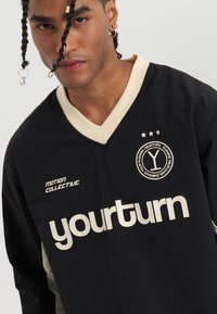 Jersey negro de manga larga con detalles en crema, presentando un cuello en V, un audaz logo "yourturn" y un emblema circular en el pecho.