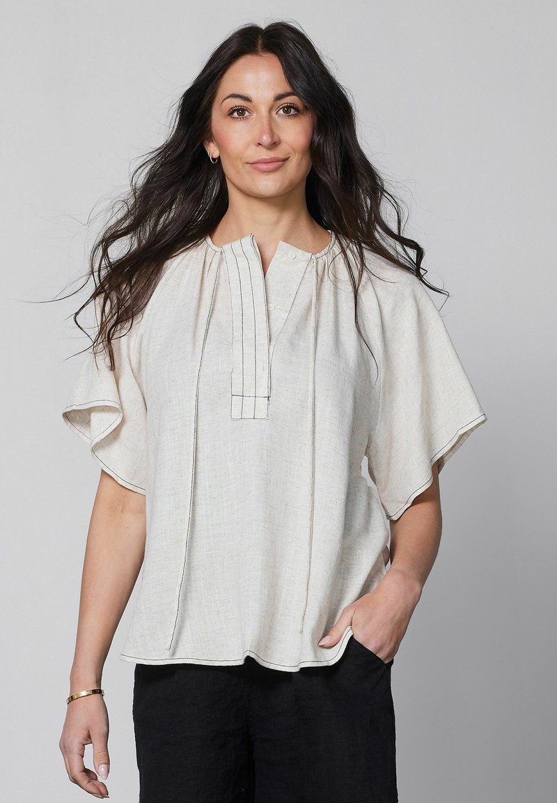 Femme aux longs cheveux foncés portant un chemisier ample beige clair à manches volantées et un pantalon noir, debout devant un fond gris uni.