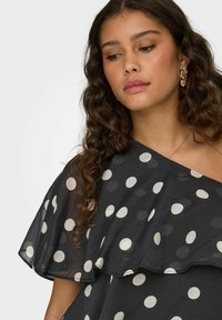 Femme aux longs cheveux bouclés portant une robe noire à une seule épaule avec de grands pois blancs et des boucles d'oreilles en forme d'anneaux torsadés dorés.