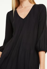 Schwarzes plissiertes Kleid mit V-Ausschnitt, langen Puffärmeln und fließender Textur. Aus leichtem Stoff mit glatter Oberfläche.