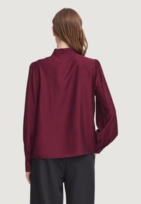 Blusa bordeaux a maniche lunghe con colletto arricciato, tessuto morbido, vestibilità rilassata e polsini con bottoni, abbinata a pantaloni scuri.