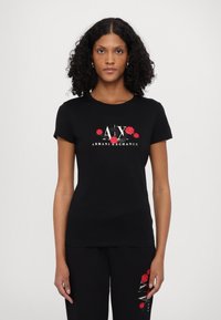 Melns īsajām piedurknēm T-krekls ar ziedu apdruku, kurā redzami sarkani ziedi un baltiem un zelta burtiem rakstīts "A|X ARMANI EXCHANGE".