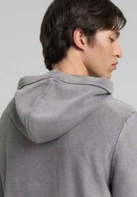 Sudadera gris con capucha, textura acanalada y corte holgado. Presenta detalles de costuras a lo largo de las uniones y una capucha con cordón ajustable.