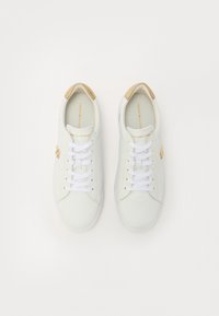 Baskets en cuir blanc avec un design à lacets, accents dorés sur le talon et le logo, tige texturée, et forme à bout rond. L'intérieur est de couleur claire.