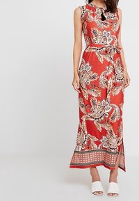 Abito maxi rosso senza maniche con un intricato motivo paisley, colletto a strati, vita con cintura e una fessura laterale all'orlo.