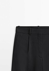 Pantalon noir ajusté avec passants pour ceinture, poches latérales et pli devant, montré de la taille jusqu'au milieu de la cuisse sur fond blanc.
