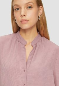 Helles lila Bluse mit Stehkragen, Knopfleiste und strukturierter Stoff mit dezenten Falten an der Halslinie.