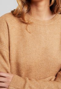 Pull beige en maille douce et texturée avec un col rond et des manches longues. Poignets et ourlet côtelés pour un ajustement confortable.