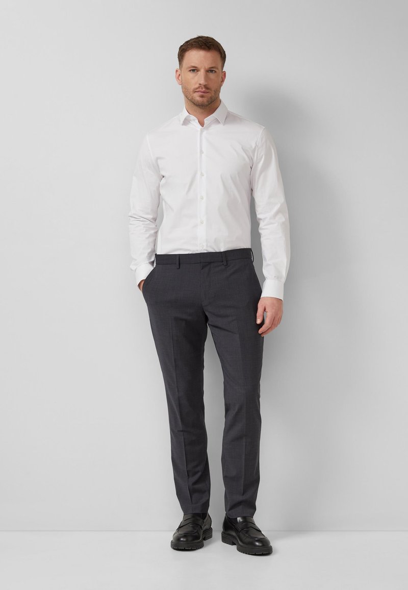 Homme debout contre un mur, portant une chemise blanche, un pantalon gris foncé et des chaussures de ville noires, les mains dans les poches.