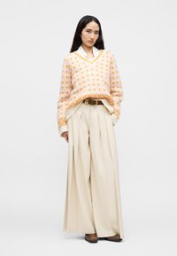 Πλεκτό πουλόβερ σε ανοιχτό ροζ με πορτοκαλί σχέδια, συνδυασμένο με μπεζ πλισέ παντελόνια με ψηλή μέση και καφέ μποτάκια μέχρι τον αστράγαλο, με λευκό γιακά.