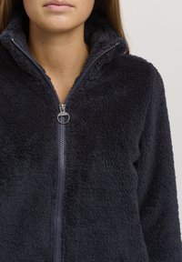 Fleece zip-up jakke i marineblå. Har høy krage, myk tekstur og en sølvglidelås med en sirkulær dragtab. Ingen mønstre eller grafikk.