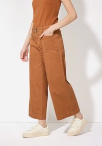 Braune weit geschnittene Hose mit zwei vorderen Taschen, hoher Taille und doppeltem Knopfverschluss. Kombiniert mit cremefarbenen Plateauschuhen.