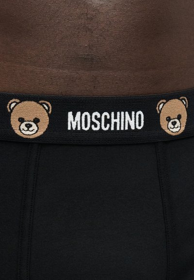 Μαύρη ζώνη εσωρούχου με το κείμενο "MOSCHINO" και δύο πρόσωπα αρκουδάκια σε σκουρόχρωμο φόντο μέσης.