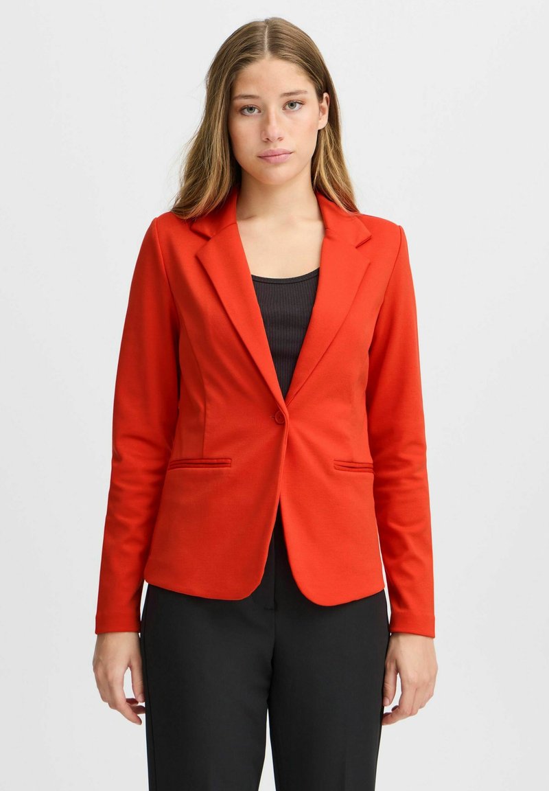 Blazer rouge avec revers en pointe, fermeture à un bouton et deux poches avant. Tissu lisse, design ajusté, associé à un haut noir et un pantalon.