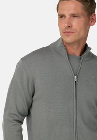 Světlé šedé svetr s zapínáním na zip a stojáčkem, ribbovanou texturou a přiléhavým střihem. Obsahuje plné zapínání na zip a bezšvová ramena.