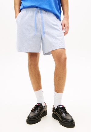 Mann iført lyseblå shorts med snor, blå skjorte, hvite ankelsokker og svarte snøresko, stående på hvit bakgrunn.