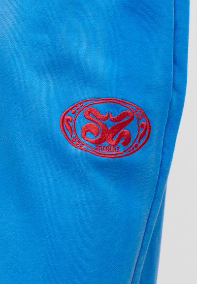 Μπλε ύφασμα με μικρό κόκκινο κεντημένο λογότυπο με αφηρημένα σχήματα μέσα σε οβάλ περίγραμμα.