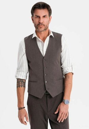 Hombre con cabello corto y oscuro y barba, vistiendo una camisa blanca de vestir con las mangas arremangadas, un chaleco gris abotonado y pantalones, mostrando tatuajes y un reloj.