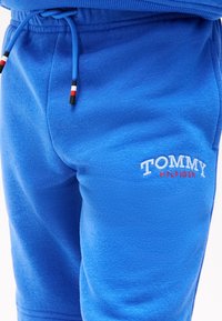 Κοντινή λήψη μπλε σορτς φούτερ με κορδόνι και λευκή και κόκκινη κεντητή επιγραφή "Tommy Hilfiger" στον μηρό.