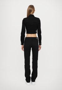 Black cropped jacket with long sleeves and a pointed collar, paired with black fitted pants and white sneakers, viewed from the back.