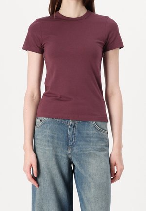 Kobieta ubrana w dopasowany, krótki, bordowy t-shirt i luźne jasnoniebieskie dżinsy na jednolitym białym tle.