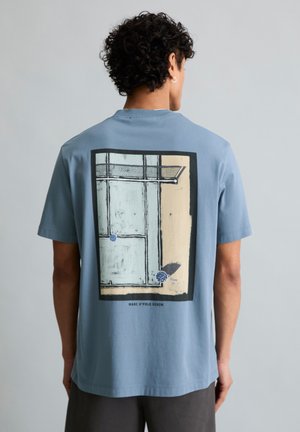 Mann trägt blaues T-Shirt mit abstraktem rechteckigem Grafikdruck und dem Schriftzug "MARC O'POLO DENIM" auf dem Rücken, steht vor einem einfarbigen grauen Hintergrund.